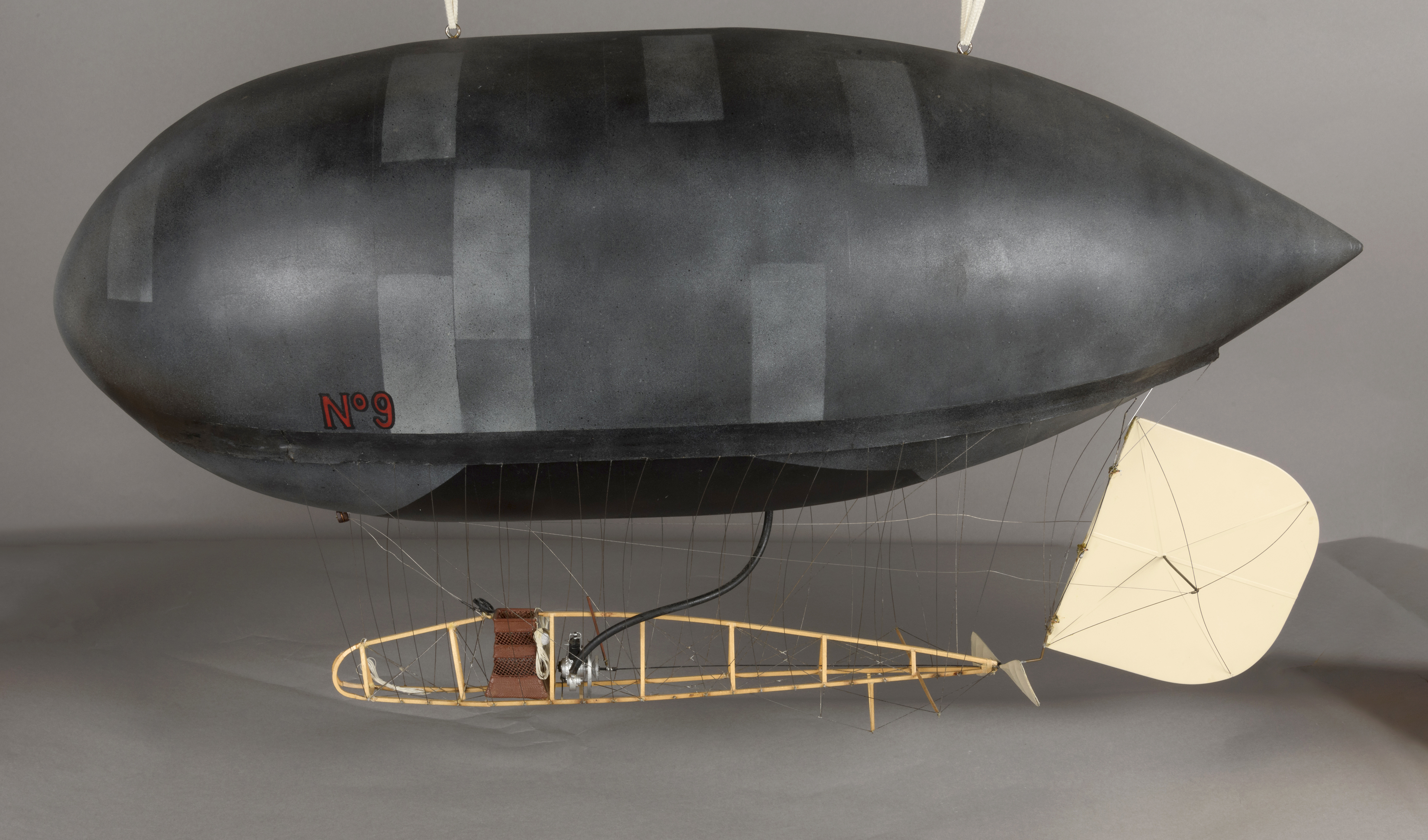 A small dark-gray airship model marked No. 9 with an exposed wooden gondola, wires, propeller, and a fabric rudder.