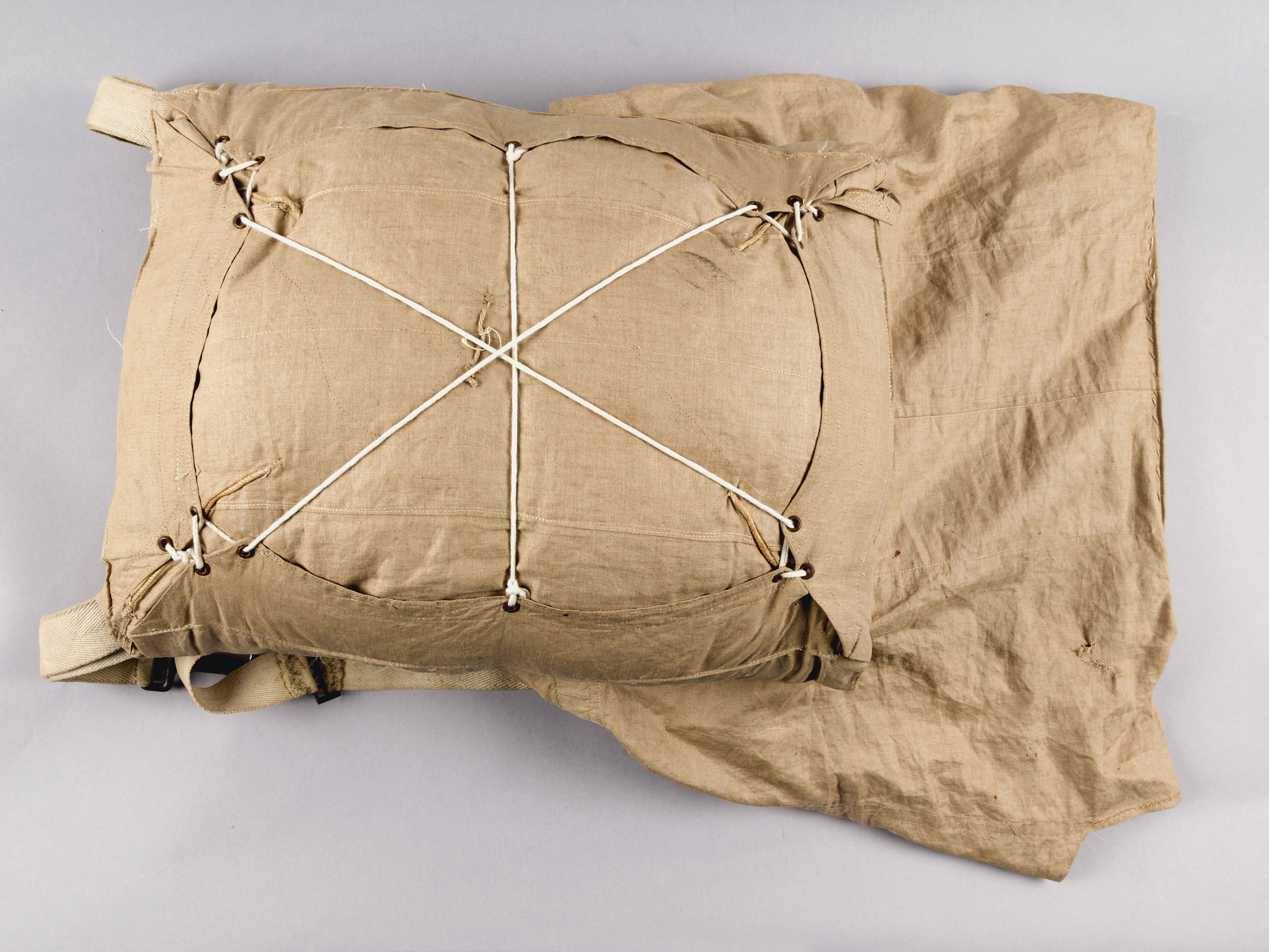 A compact cream-colored parachute pack tied with light cord in a crossed pattern.