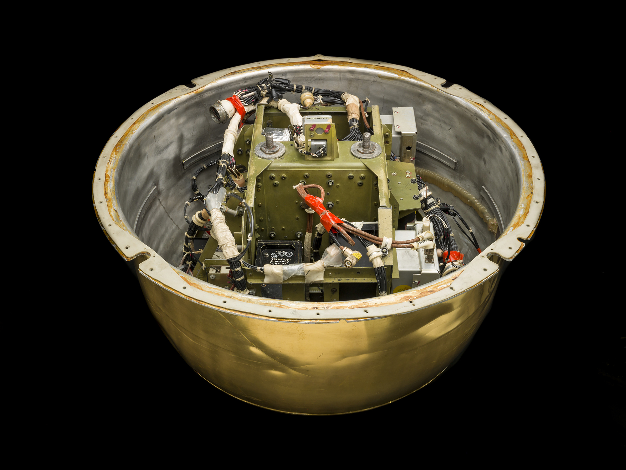 A circular reentry capsule assembly with dense wiring and components inside a perforated gold-colored casing.