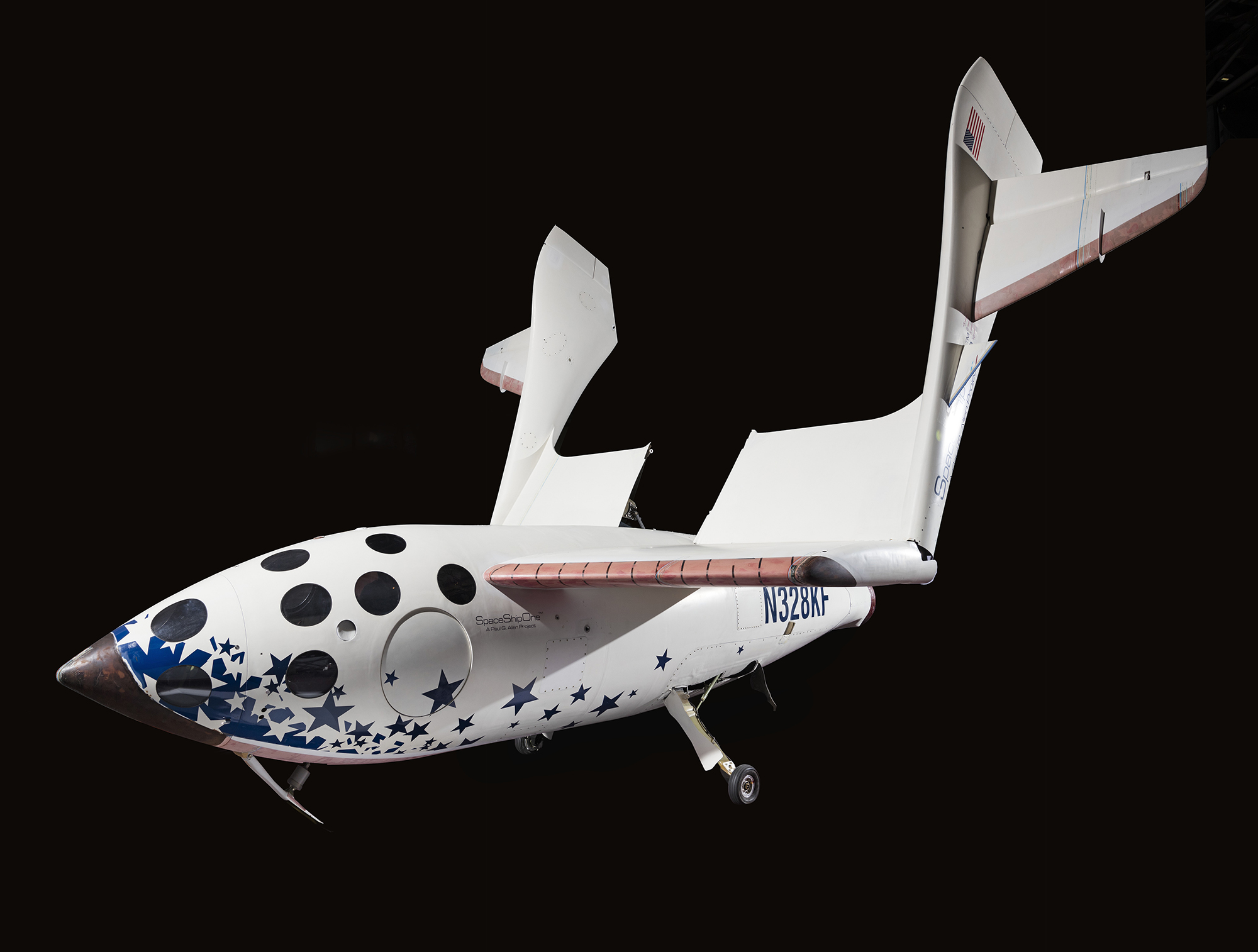A white piloted spaceplane with a cigar-shaped fuselage, twin tail booms, blue stars around the nose, red trim, circular windows, and a feathering wing system.