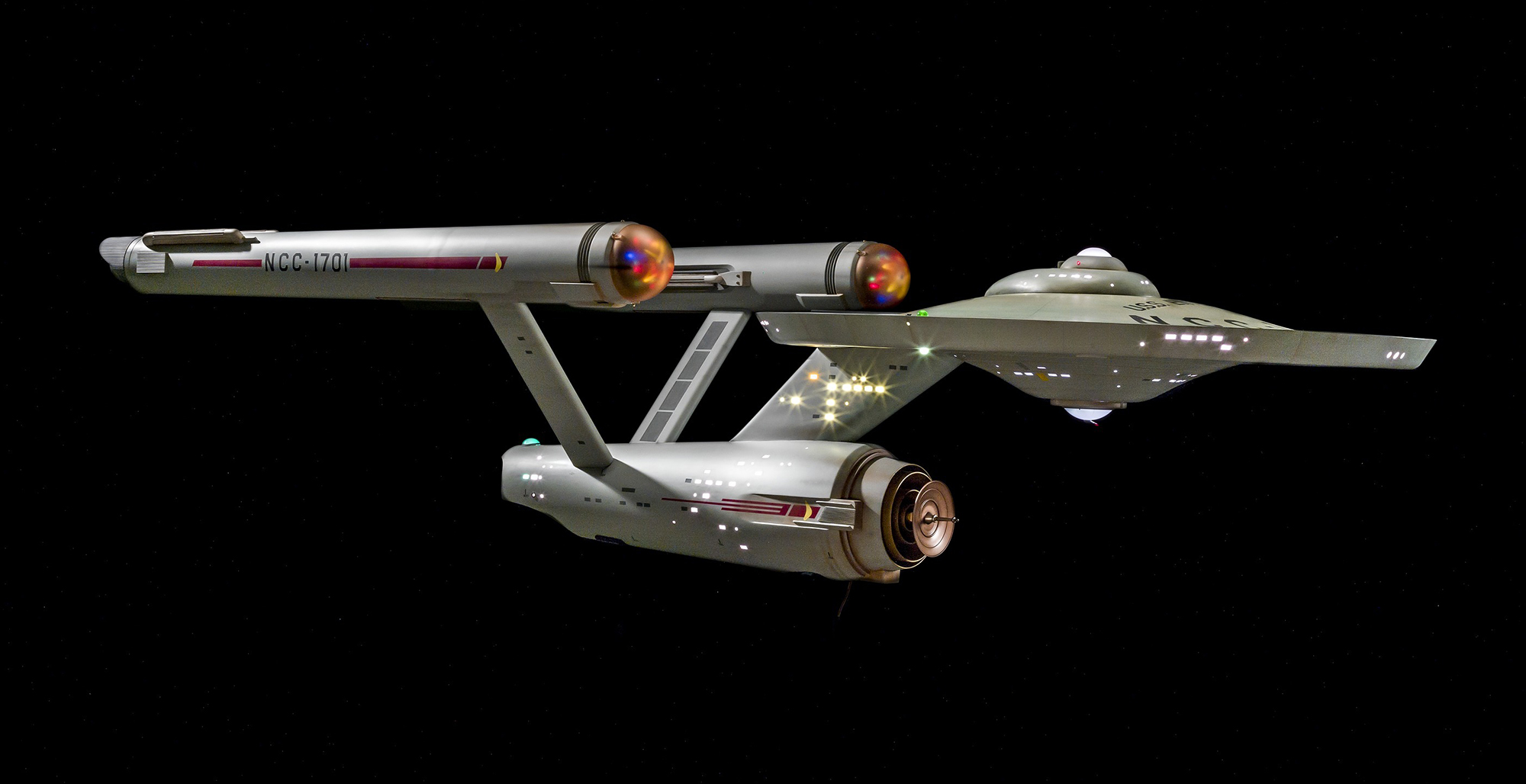 A large gray television studio model with a saucer-shaped forward section, a central hull, and two long engine nacelles, detailed and lettered mainly on the side filmed by the camera.