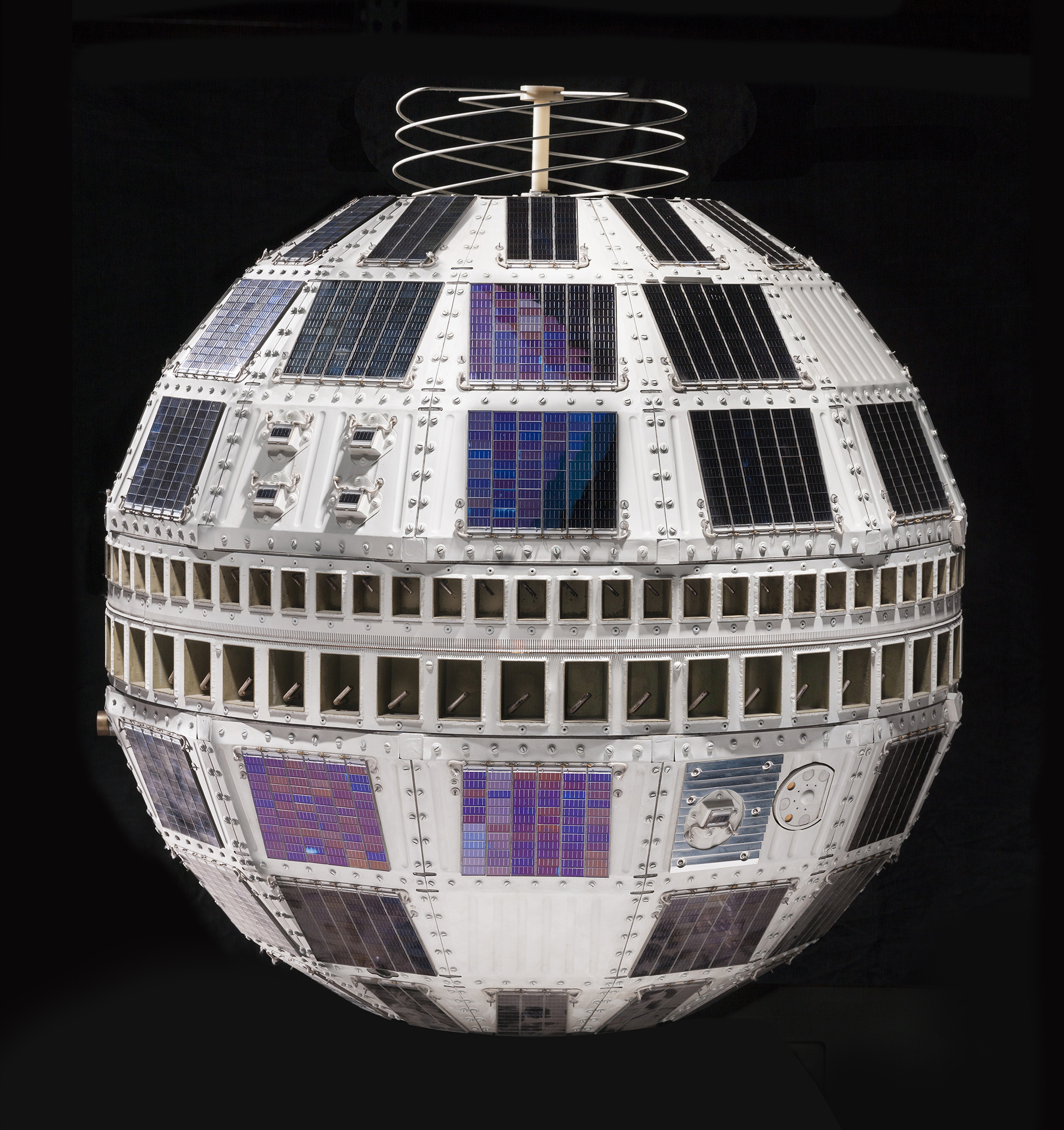 A spherical communications satellite covered with rows of black solar cells, narrow center bands, and a spiral antenna on top.
