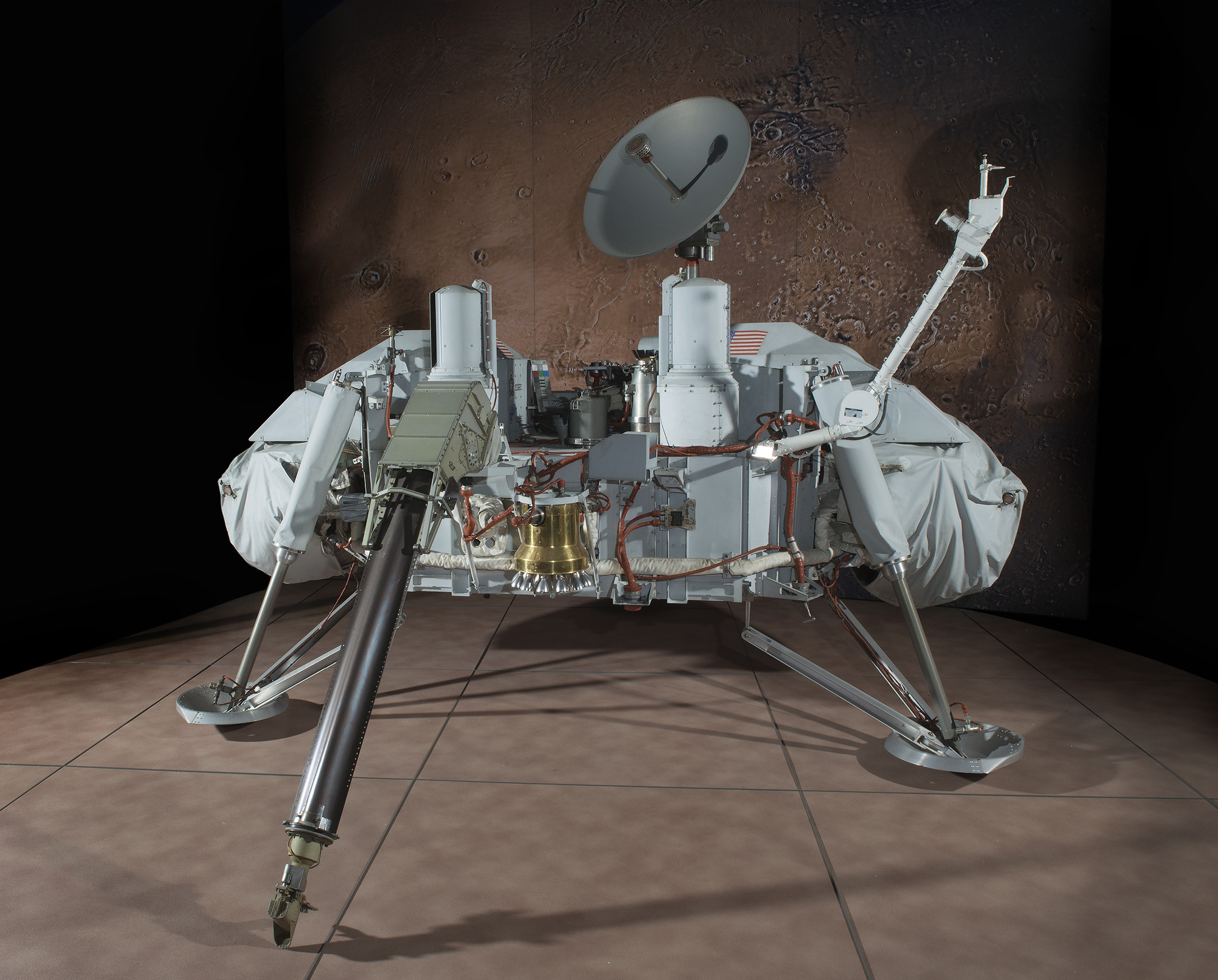 A three-legged Mars lander with a white instrument body, broad footpads, two cylindrical cameras, a sampling arm, and a circular high-gain antenna.