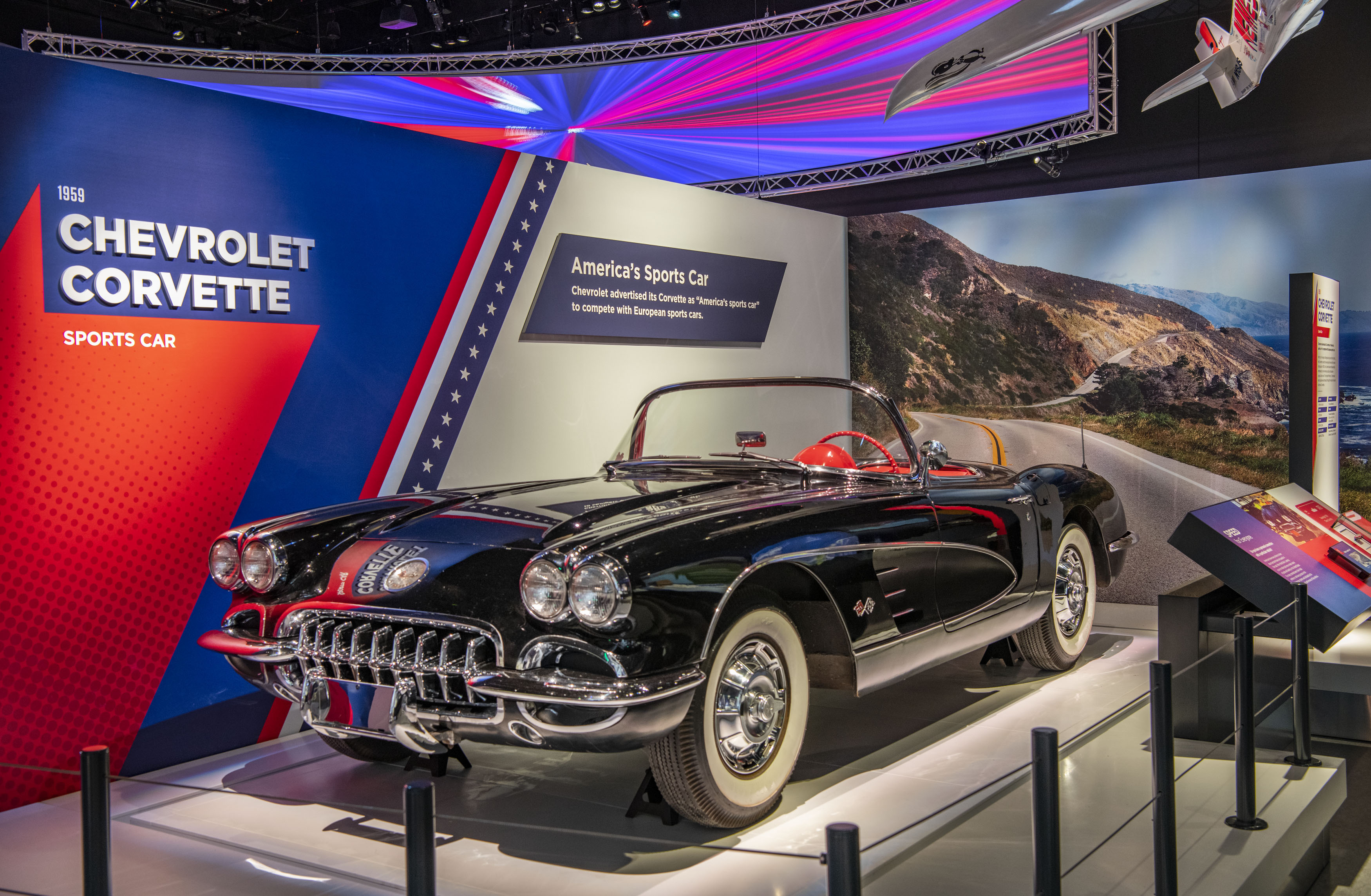 Black two-seat convertible sports car with red interior, whitewall tires, bright side trim, and a low windshield.