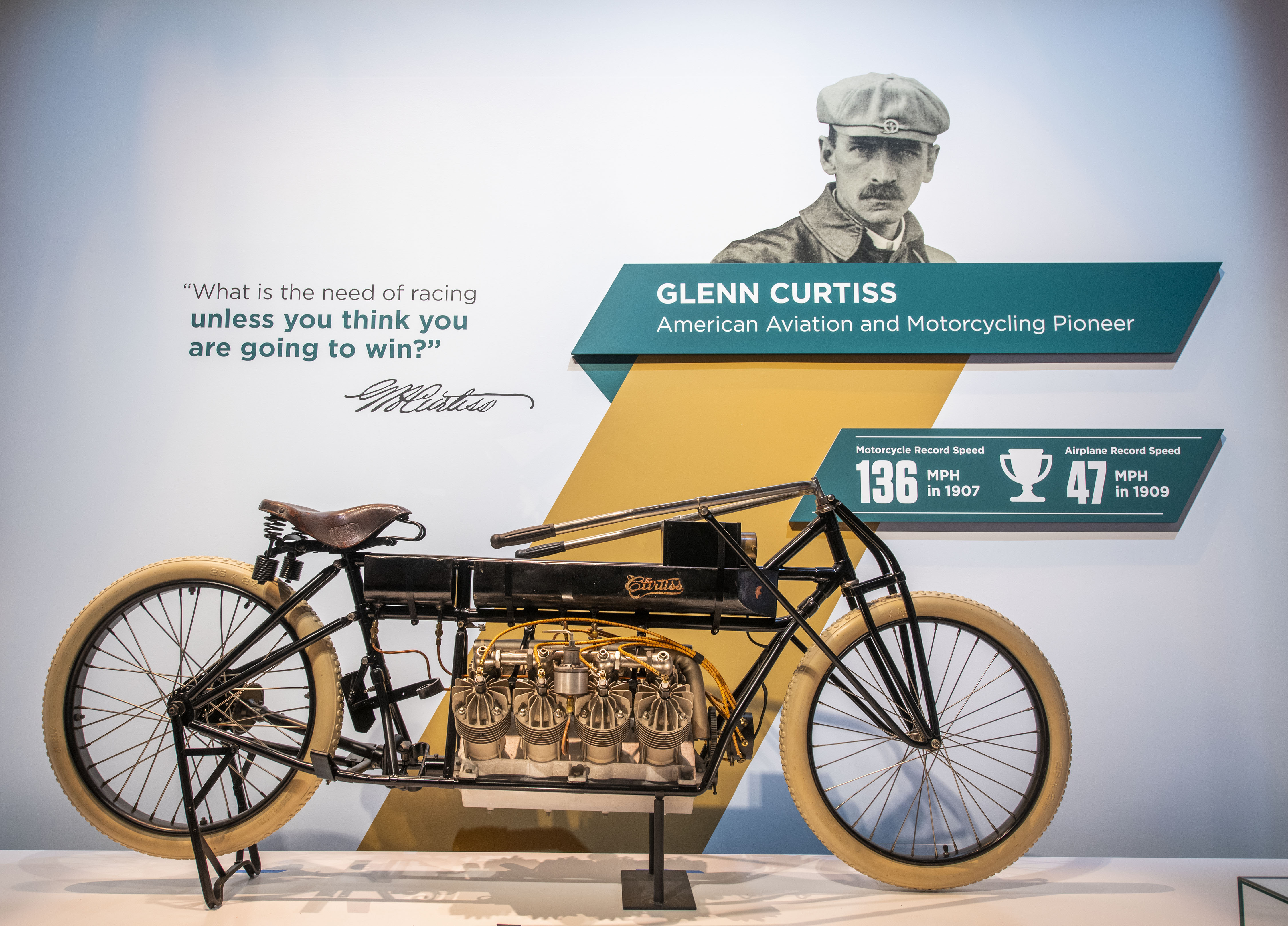 Black open-frame motorcycle with white tires, brown saddle, low handlebars, and a large exposed Curtiss V-8 engine.