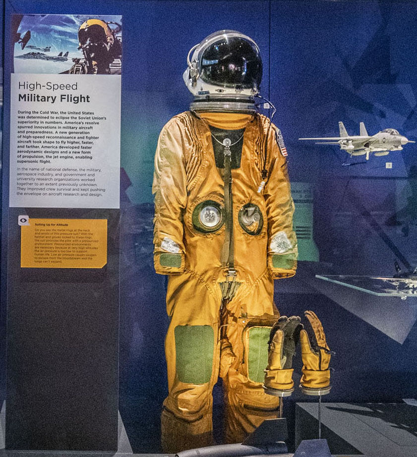 Yellow full-pressure flight suit with dark closures and knee pads, yellow gloves, and a white helmet with a dark visor.