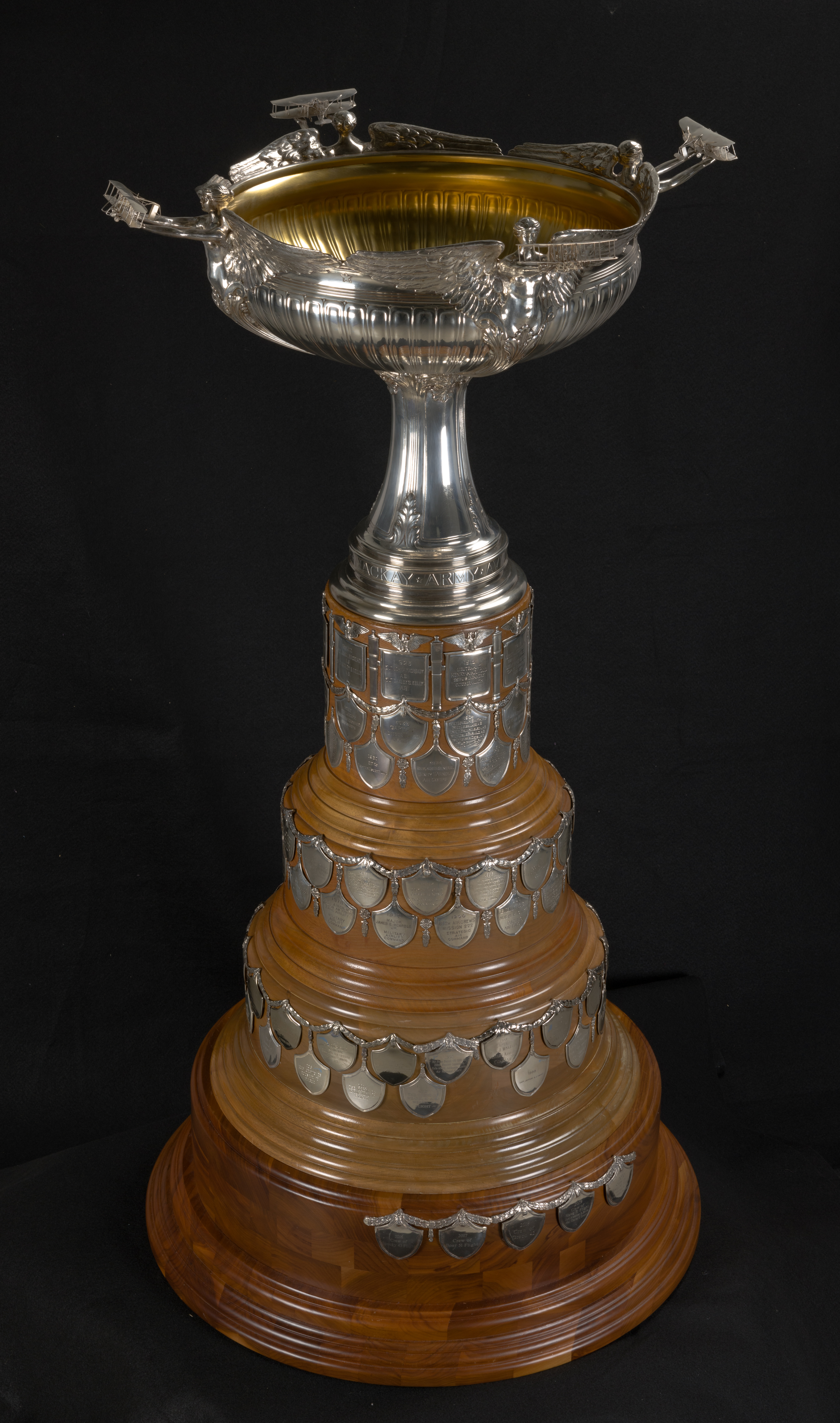 Silver Mackay Trophy with four winged figures supporting a gold-lined cup on an inscribed wooden base.